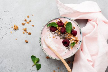 Pembe ahududu muz smoothie kaseyle granola, chia tohum, Hindistan cevizi ve ışık gri beton zemin üzerine nane. Seçici odak. Üstten Görünüm. Kopya alanı.