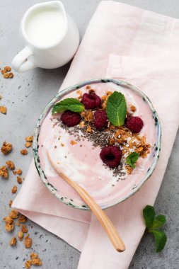 Pembe ahududu muz smoothie kaseyle granola, chia tohum, Hindistan cevizi ve ışık gri beton zemin üzerine nane. Seçici odak. Üstten Görünüm. Kopya alanı.