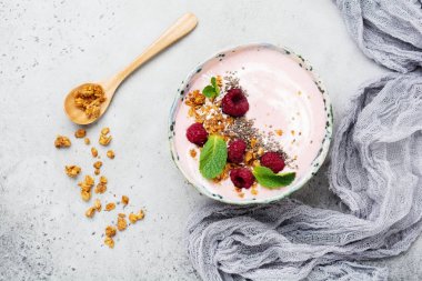 Pembe ahududu muz smoothie kaseyle granola, chia tohum, Hindistan cevizi ve ışık gri beton zemin üzerine nane. Seçici odak. Üstten Görünüm. Kopya alanı.