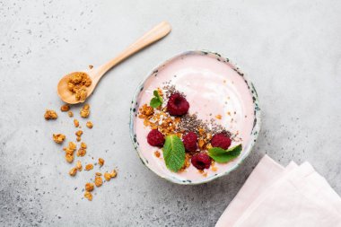 Pembe ahududu muz smoothie kaseyle granola, chia tohum, Hindistan cevizi ve ışık gri beton zemin üzerine nane. Seçici odak. Üstten Görünüm. Kopya alanı.