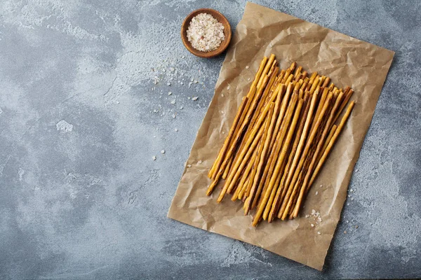 Salted bread sticks or long crunchy salty pretzel sticks on parchment paper on old gray stone or concrete background. Selective focus. Top view.