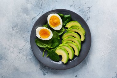 Sağlıklı vejetaryen salata kahvaltısı. Ispanak yaprakları, domates, avokado ve gri tabakta haşlanmış yumurta ve gri beton arka plan. Buda kasesi. Üst görünüm. 