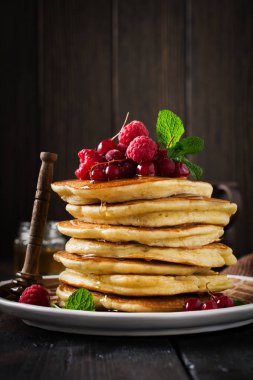 Eski bir ahşap arka plan üzerinde bal, taze ahududu ve kırmızı kuş üzümü ile ev yapımı küçük krep yığını. Seçici odaklama.