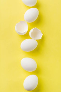Beyaz çiğ tavuk yumurtaları dikey sırada yatıyor ve sarı arka planda kırık yumurta var. Üst görünüm.