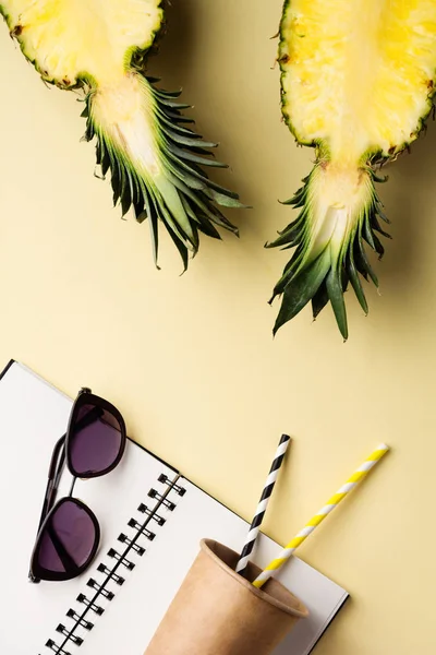 Fresh pineapple cut in two part, notebook or sketchbook and sunglasses, paper cup for cocktail on yellow background. Summer concept. Creative flat lay with copy space. Top view.