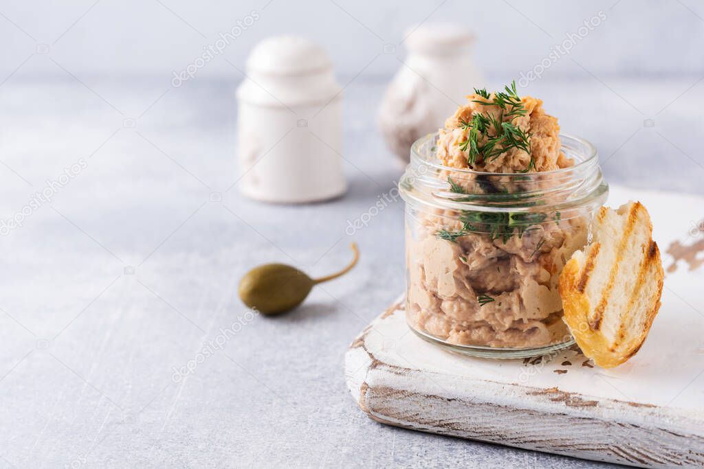 Chicken meat paste pate in a jar served with capers on a light concrete background.