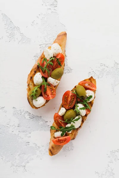 Caprese bruschetta toasts with cherry tomatoes, mozzarella, olives and basil on white plate old light background. Top view.