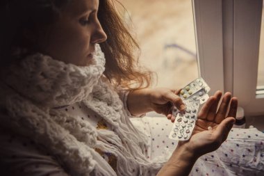 Genç bir kız hastalandı ve elinde haplarla pencerenin kenarında oturuyordu. Hasta kadın elinde hap tutuyor..