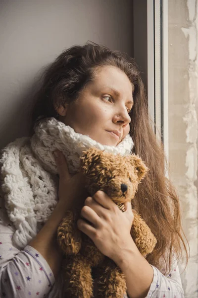 Sick girl sitting with a bear in the hands of the window. Young girl is sick and stays at home by a window with a scarf and teddy bear.