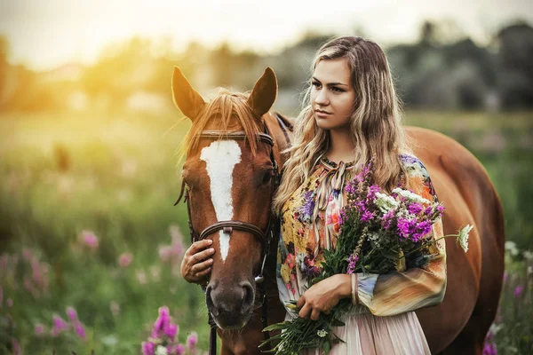 Jovem mulher fica perto de um cavalo em um vestido de verão em um prado — Fotografia de Stock