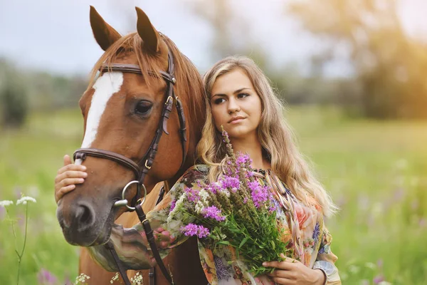 Mujer joven se para cerca de un caballo en un vestido de verano en un prado — Foto de Stock