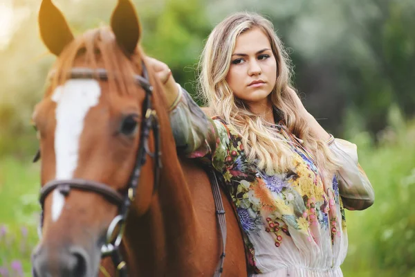 Mujer joven se para cerca de un caballo en un vestido de verano en un prado — Foto de Stock