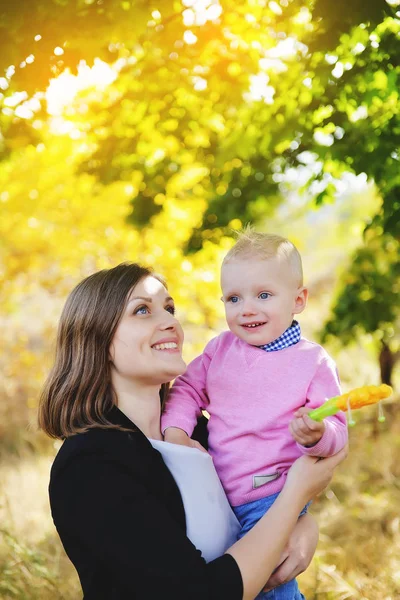 Uma jovem mãe segurando um pequeno filho em seus braços e de pé em um parque perto do bordo amarelo no outono . — Fotografia de Stock