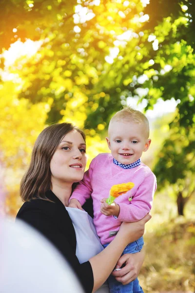 Een jonge moeder houden een klein zoon in haar armen en permanent in een park in de buurt van de gele maple in de herfst. — Stockfoto