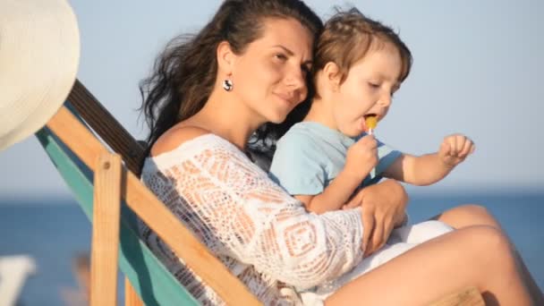 Anya ül a fiával, egy karosszékben a tengerparton napnyugtakor nyáron. — Stock videók