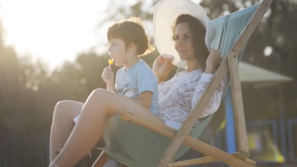 Moeder zit met haar zoon in een fauteuil op het strand in de zomer bij zonsondergang. — Stockvideo