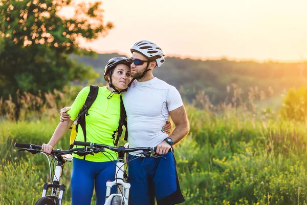 Un hombre y una mujer en bicicleta se paran juntos en un parque cerca de sus bicicletas en la primavera al atardecer. El hombre abrazó a la mujer . — Foto de Stock