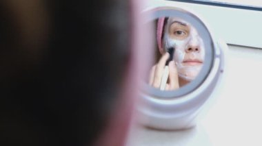 Kadın yüzünü bir fırça ile kozmetik maske koyar ve aynaya bakar.