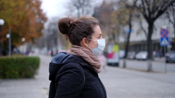 Eine junge Frau mit medizinischer Maske steht im Herbst auf einer Straße in einer Stadt. — Stockvideo