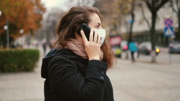 Uma jovem mulher está de pé em uma rua da cidade em densa névoa em uma máscara médica e falando ao telefone . — Vídeo de Stock