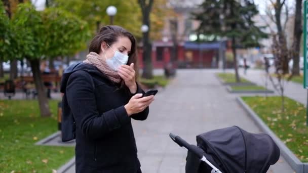 Mladá matka s kočárkem v parku v lékařské masce mluví po telefonu. — Stock video