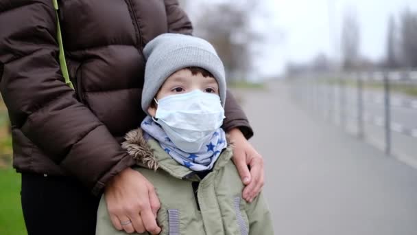 Egy kisfiú orvosi maszkban anya mellett áll az utcán.. — Stock videók