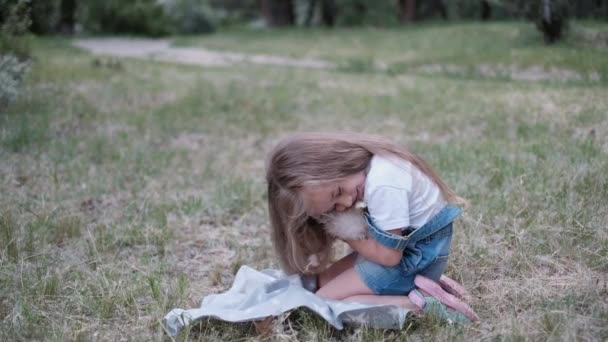 Linda niña se sienta en la hierba y sostiene un cachorro blanco en sus brazos en el verano . — Vídeo de stock