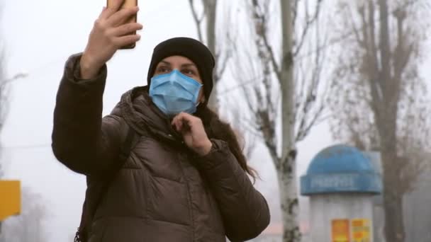 Młoda kobieta robi sobie selfie w ochronnej masce medycznej na ulicy w miejscu publicznym. Ochrona przed chińskim koronawirusem. — Wideo stockowe