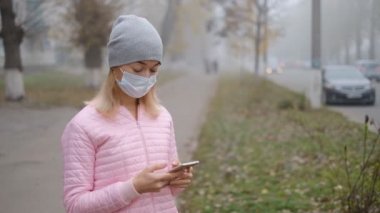 Coronavirus koruması. Tıbbi koruyucu maskeli genç bir kadın Avrupa 'da bir şehir caddesinde akıllı telefonuyla duruyor. Çin 'den gelen koronavirüsün belirtileri ortaya çıktı..