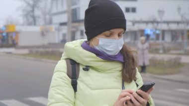 Coronavirus koruması. Tıbbi koruyucu maskeli genç bir kadın Avrupa 'da bir şehir caddesinde akıllı telefonuyla duruyor. Çin 'den gelen koronavirüsün belirtileri ortaya çıktı..