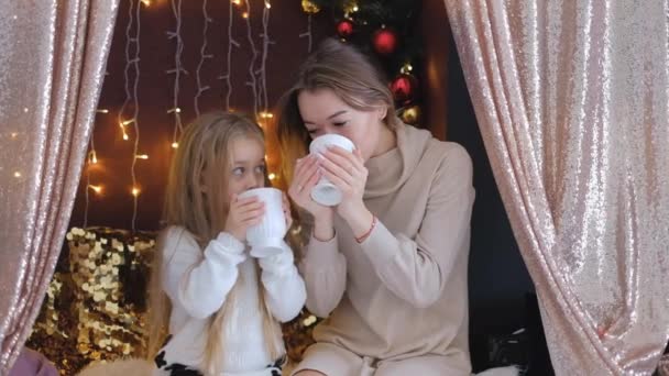 Moeder met dochter zit op een bank met kopjes warme chocolademelk in Kerstmis. — Stockvideo