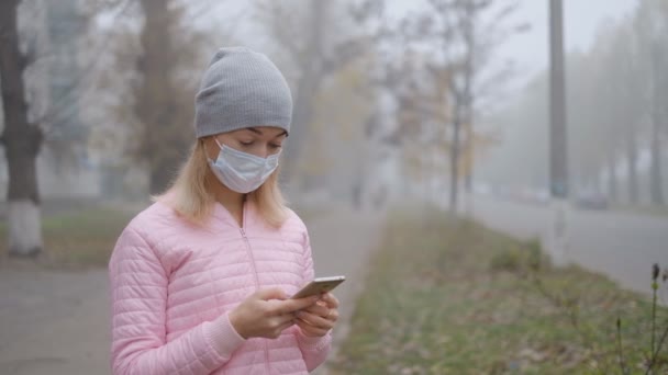 Protection contre le coronavirus. Une jeune femme portant un masque de protection médicale se tient avec un smartphone dans une rue de la ville en Europe. — Video