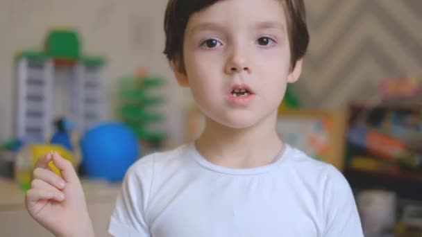 Mały chłopiec stoi w pokoju dziecięcym i potrząsa palcami jak mleczny ząb. Chłopak wyciąga mleczny ząb z ust i jest zaskoczony.. — Wideo stockowe
