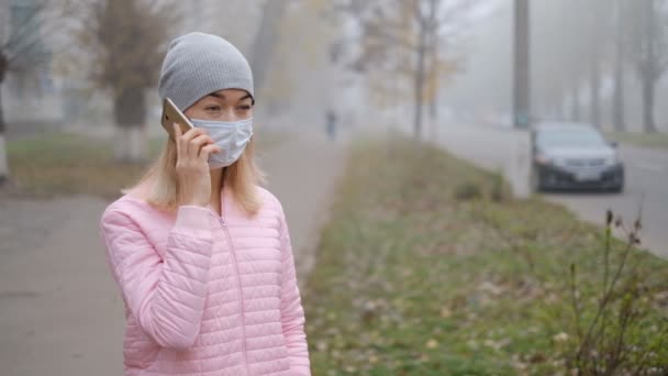 Защита от коронавируса. Молодая женщина в медицинской защитной маске стоит со смартфоном на городской улице в Европе . — стоковое видео
