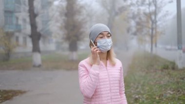 Coronavirus koruması. Tıbbi koruyucu maskeli genç bir kadın Avrupa 'da bir şehir caddesinde akıllı telefonuyla duruyor..