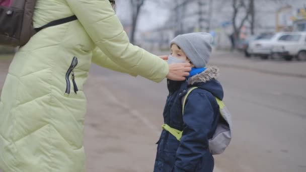 Schutz vor dem chinesischen Coronavirus in einer europäischen Stadt. Mutter korrigiert einem kleinen Sohn auf der Straße eine medizinische Schutzmaske. — Stockvideo