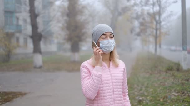 Coronavirusbescherming. Een jonge vrouw met een medisch beschermend masker staat met een smartphone op een straat in Europa. — Stockvideo