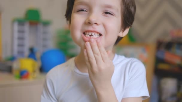 Een jongetje staat in een kinderkamer en schudt met zijn vingers een melktand. De jongen trekt een melktand uit zijn mond en is verbaasd. — Stockvideo