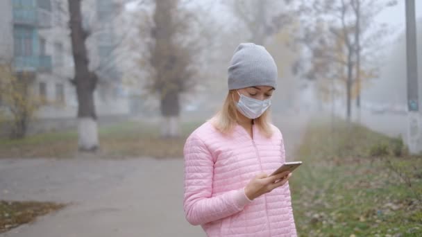 Coronavirus protection. A young woman in a medical protective mask stands with a smartphone on a city street in Europe. — Stock Video