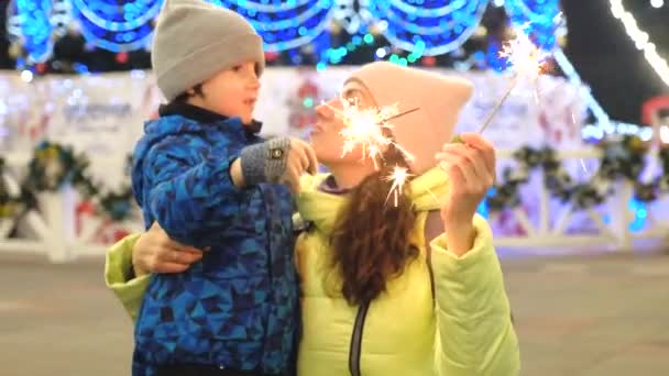 Mamá e hijo celebran las vacaciones de invierno cerca del árbol de Navidad en el mercado de Navidad. Las chispas en las manos de la mamá y el hijo por la tarde cerca del árbol de Navidad en invierno . — Vídeos de Stock