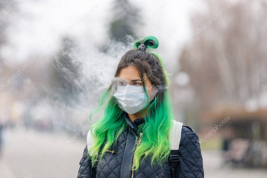 Girl with green hair in a protective medical mask and smokes a vape.