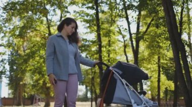 Genç bir anne bebek arabasında uyuyan bir bebekle parkta yürüyor..