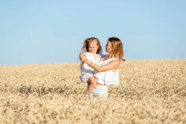 Een jonge vrouw staat in de zomer in een graanveld en houdt haar dochtertje in haar armen. — Stockfoto