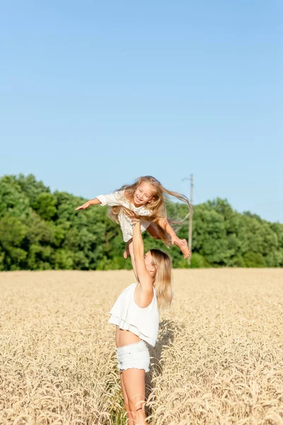 Eine junge Frau steht im Sommer in einem Weizenfeld und hält ihre kleine Tochter im Arm. — Stockfoto