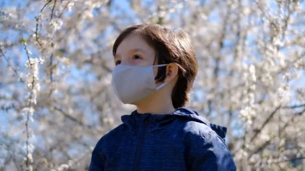 在检疫和验尸官流行病期间 一个小男孩戴着防护面罩离开了家 去散步 一个男孩戴着防护面具站在一棵开花的树旁边的公园里 — 图库视频影像