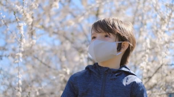 Ένα μικρό αγόρι έφυγε από το σπίτι για μια βόλτα με μια προστατευτική μάσκα κατά τη διάρκεια της καραντίνας και της επιδημίας του κορωναϊού. Ένα αγόρι στέκεται σε ένα πάρκο κοντά σε ένα ανθισμένο δέντρο με προστατευτική μάσκα.. — Αρχείο Βίντεο