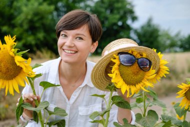 Güzel bir genç kadın yazın ayçiçeği tarlasında duruyor ve tatlı tatlı gülümsüyor..