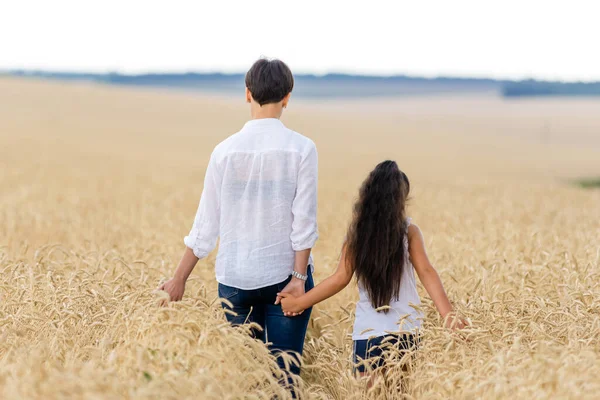 Mother and daughter joined hands and walk along a wheat field in summer. A girl with long, beautiful hair.