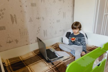 Koronavirüs salgını sırasında çocuklar için uzaktan öğrenme. Çocuk yatakta oturuyor ve interneti ve bilgisayarı kullanarak bir okul ödevi alıyor..
