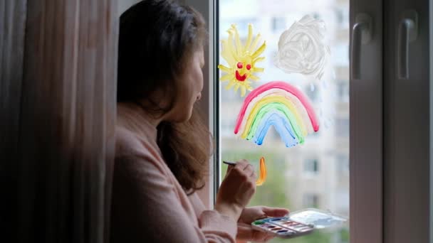 La mujer dibuja un arco iris en la ventana. Una mujer está en cuarentena en casa. — Vídeos de Stock